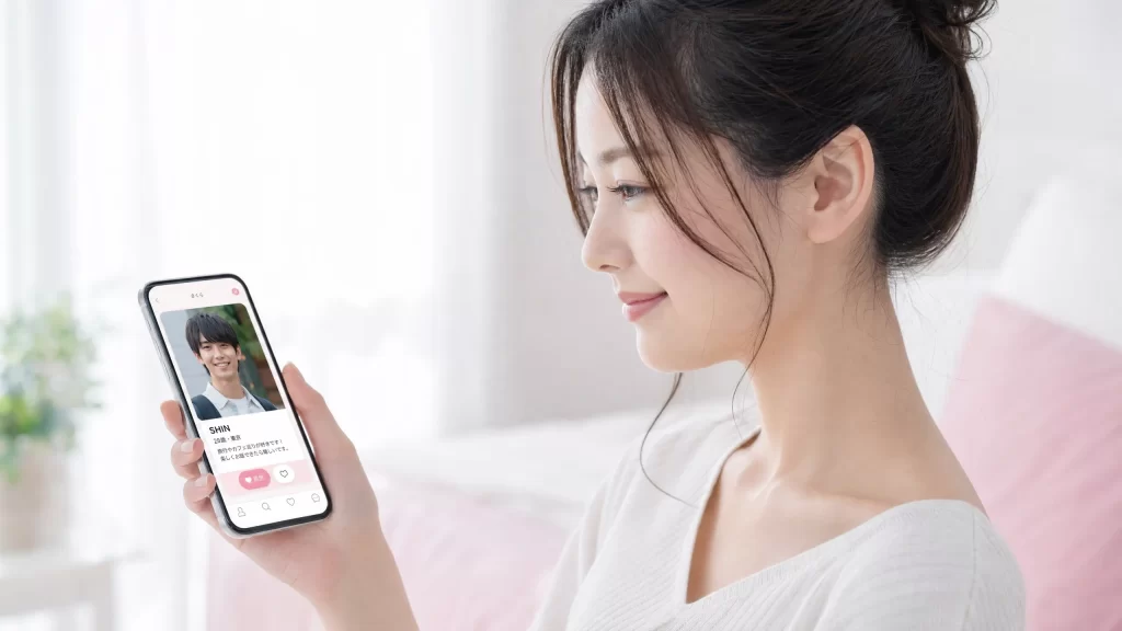 スマートフォンを手に持った女性が画面を見ている横顔、画面にはマッチングアプリ風のシンプルなUIが映る、明るく前向きな雰囲気、ピンクとホワイトの優しい配色