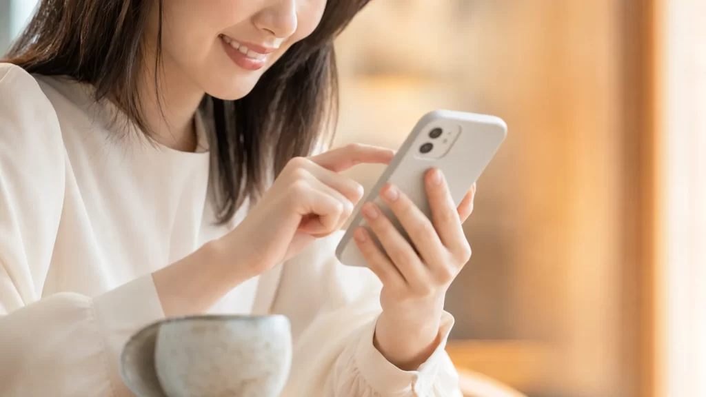 カフェのテーブルで微笑みながらスマートフォンを操作する女性の手元と顔の下半分、温かいベージュとオレンジの配色、優しく前向きな雰囲気