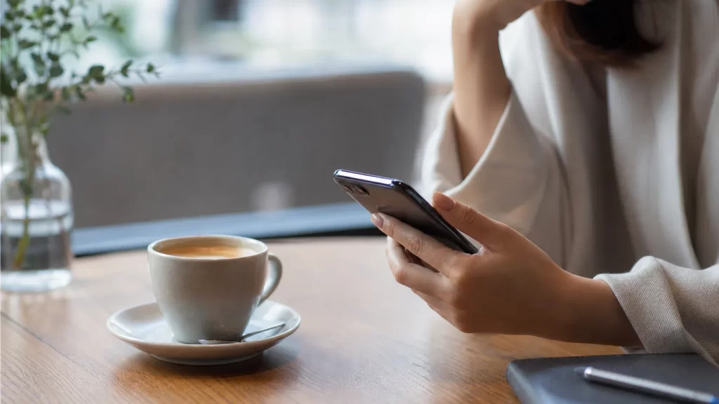 都心のおしゃれなカフェのテーブルに置かれたコーヒーカップと、女性の手元のスマートフォン。